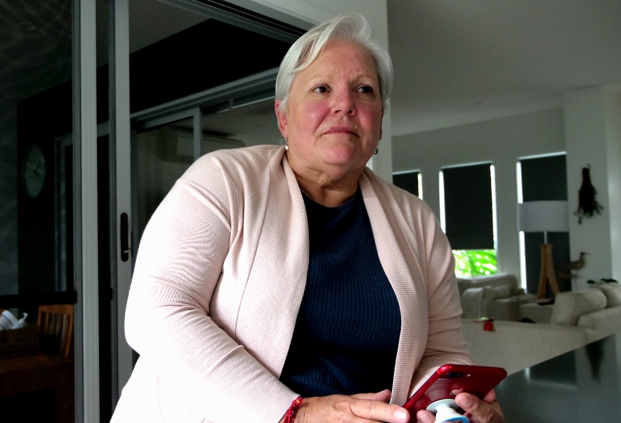 A woman with white hair stands at the kitchen bench with her phone in hand
