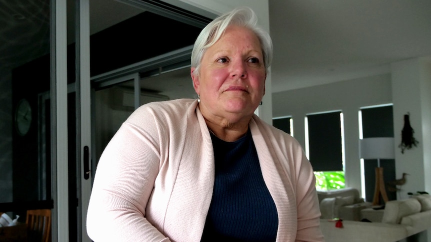 A sombre woman with white hair stands at the kitchen bench with her phone in hand, pink cardigan, blue top.