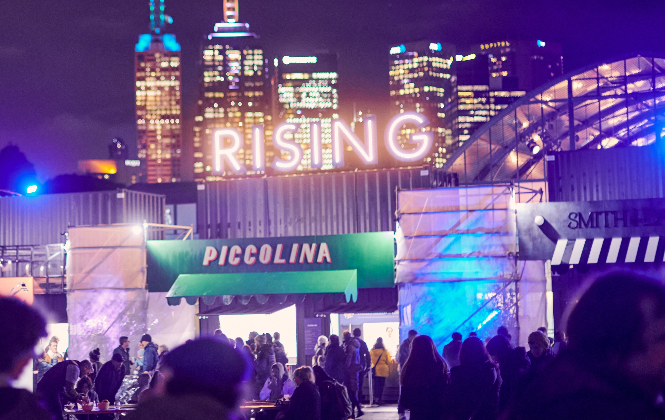 A sign reads RISING above the entrance to a live music venue and bar and food stalls, the Melbourne skyline visible behind 