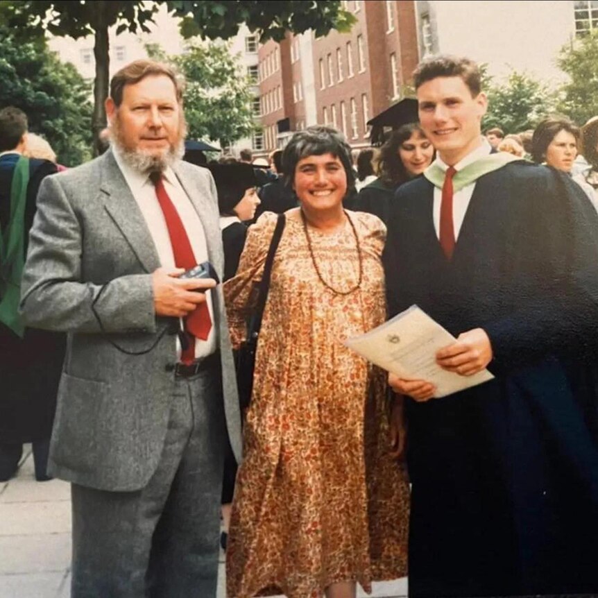 Two men and a woman smile at the camera. One of the men is wearing academic regalia.