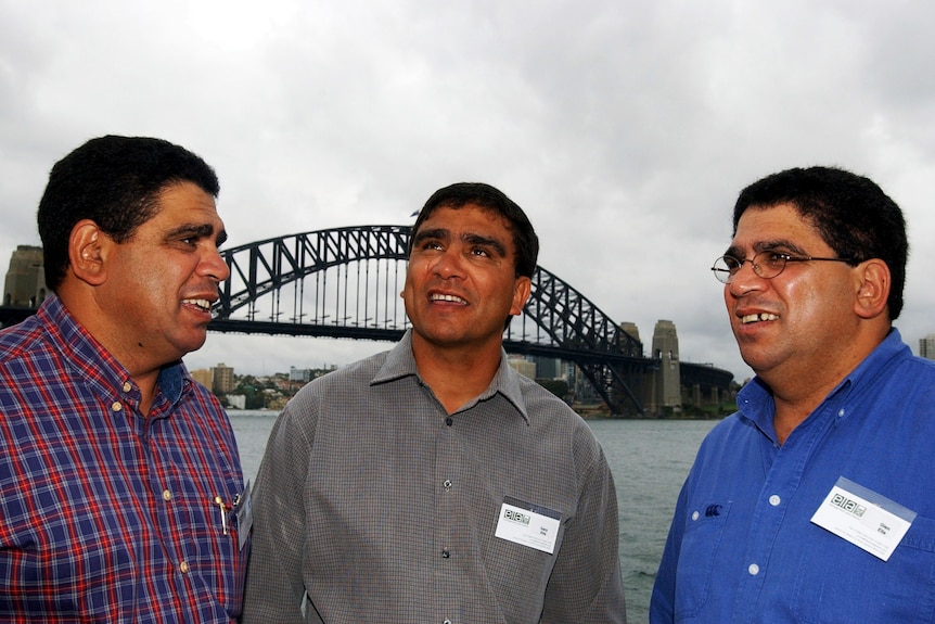 Mark, Glen and Gary Ella stand together in front of the Sydney Harbour Bridge