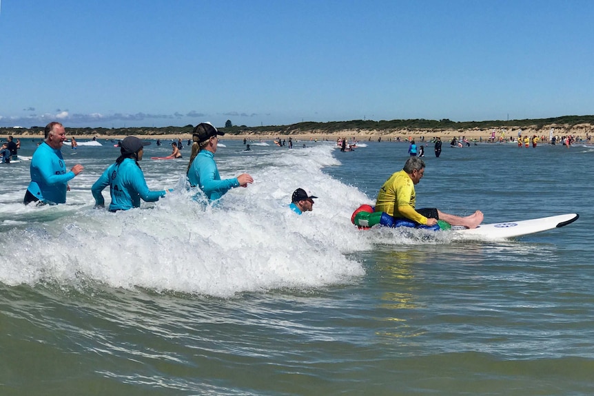 At a surf beach, a man sitting on a large surf board as it's pushed by the waves. Helpers look on.