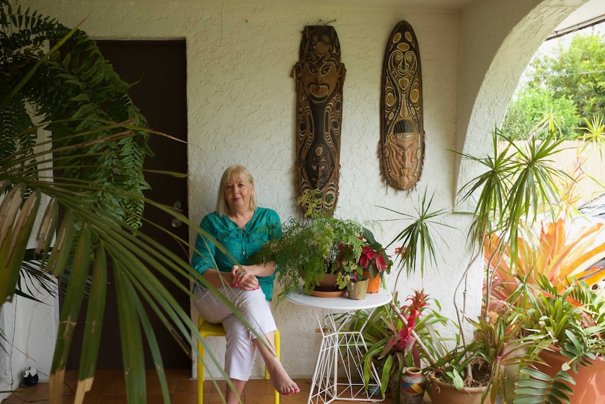 A woman with length blonde hair and a green blouse sits amongst plants and Pacific Islander art in her home.