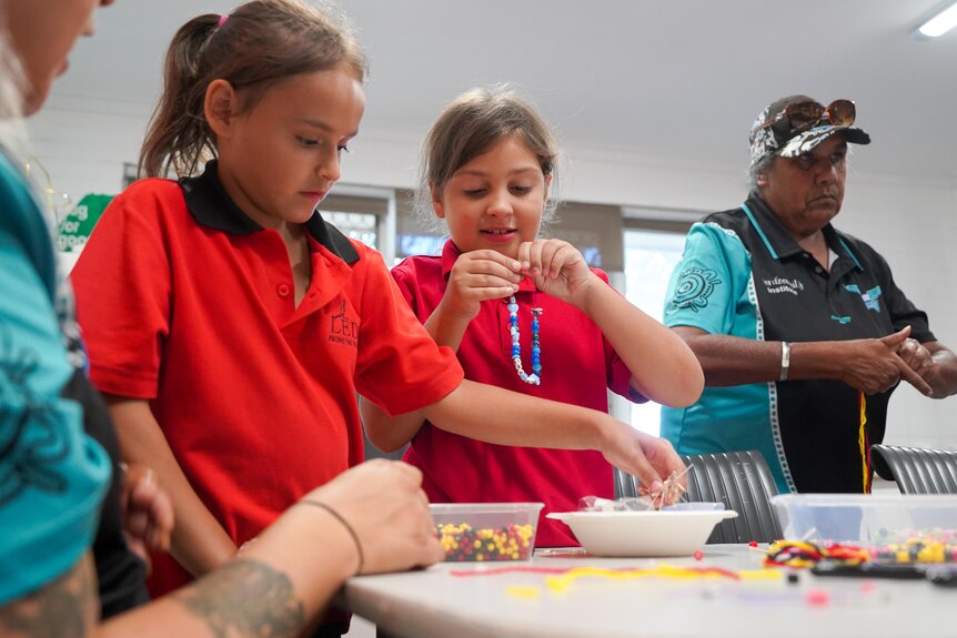Two girls wearing red school uniform shirts are playing with beads. An Indigenous elder in the background with thread in hand. 
