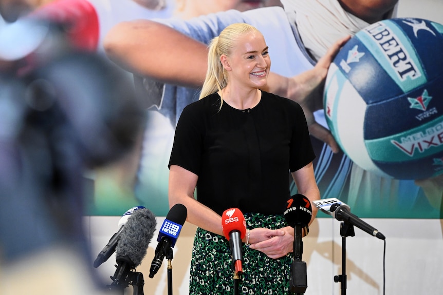 A woman stands in front of several microphones and smiles as she listens