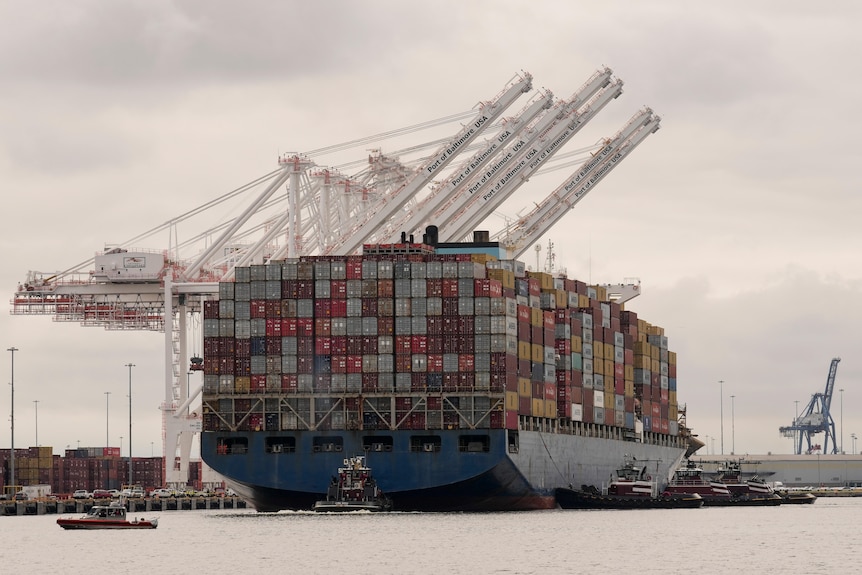 A large ship with containers on board with cranes above and tugboats next to it.