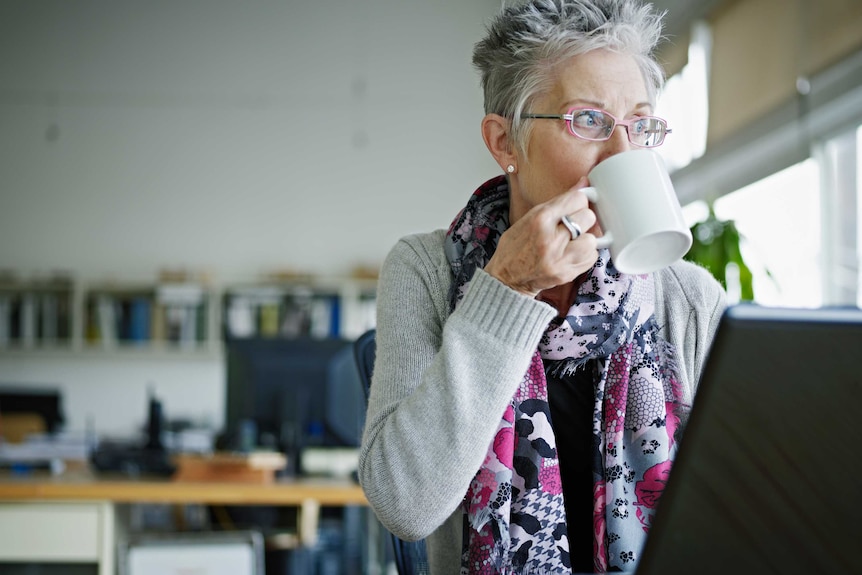 In a room with a blurred background, a woman sits in front of a laptop with a mug to her mouth looking sideways towards window.