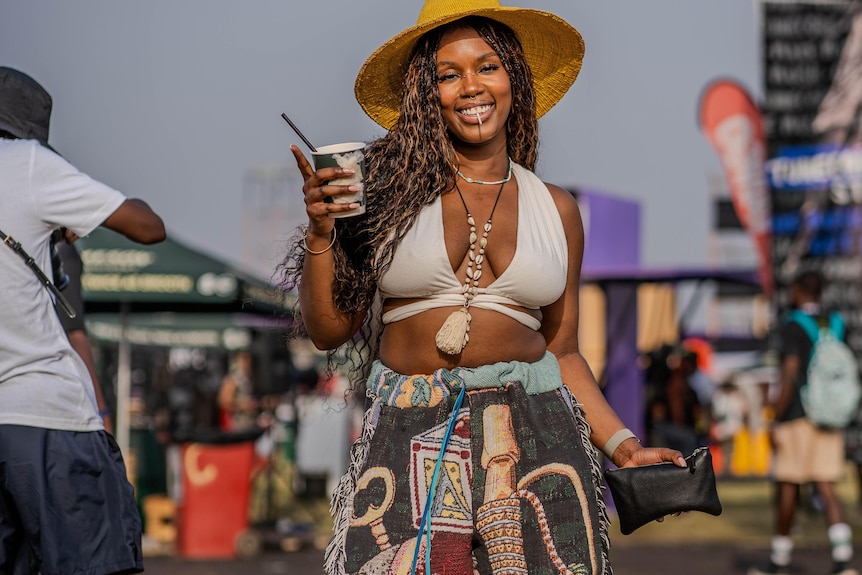 A woman holding a cup, wearing a hat, smiling, with a crop top and colourful pants