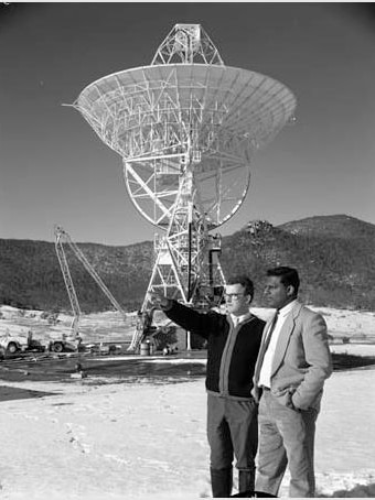 Two men stand in snow in front of radio telescope