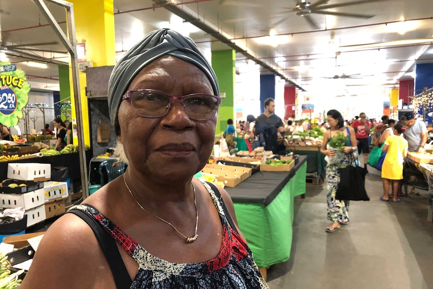 Papua New Guinean woman in blue turban stands in front of indoor market stalls.