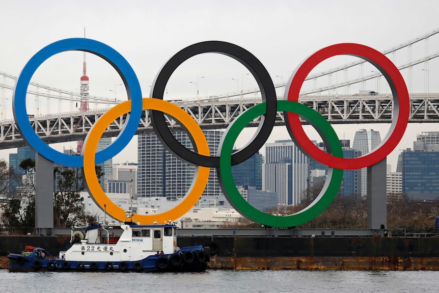 Giant versions of the five-coloured Olympic rings stand in front of a view of the city of Tokyo.