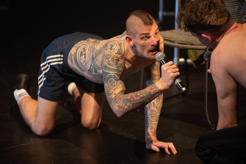 A shirtless man with tattoos on all fours on a stage floor, speaking into a microphone as another man watches on.