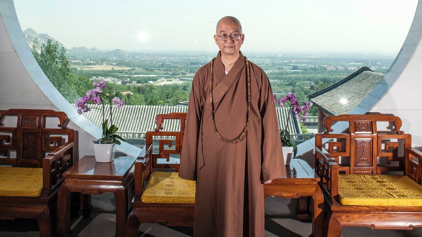 Abbot Xuecheng of the Beijing Longquan Temple. He is posing in front of a window wearing a brown robe and glasses.