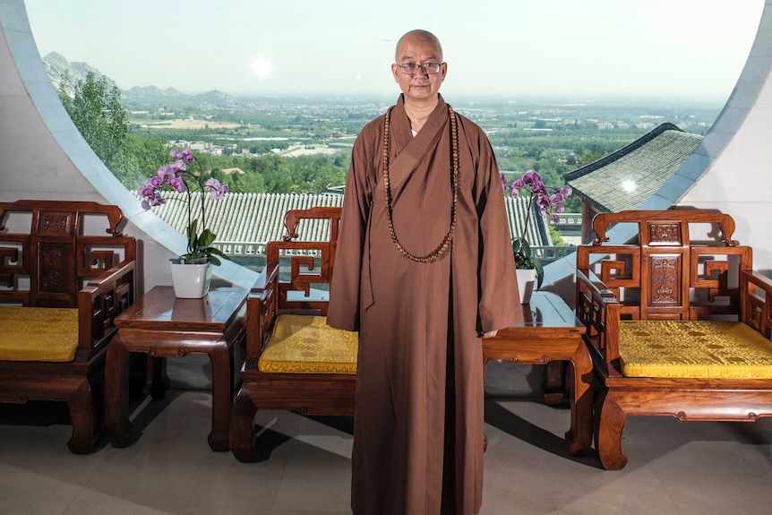 Abbot Xuecheng of the Beijing Longquan Temple. He is posing in front of a window wearing a brown robe and glasses.