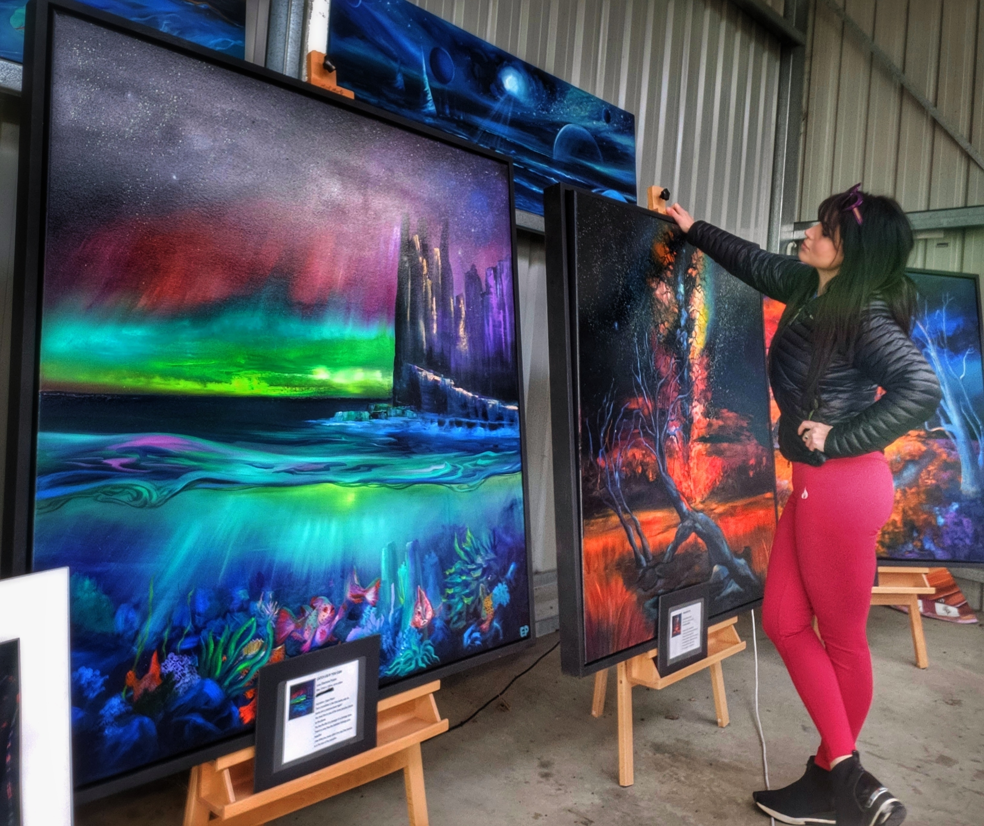 A brunette woman in pink tights stands and looks at her large landscape paintings