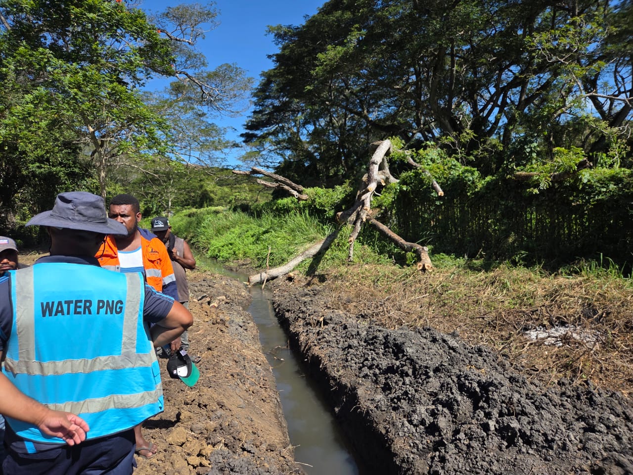 A group of Water PNG workers stand next to a trench that has been dug in rugged terrain.