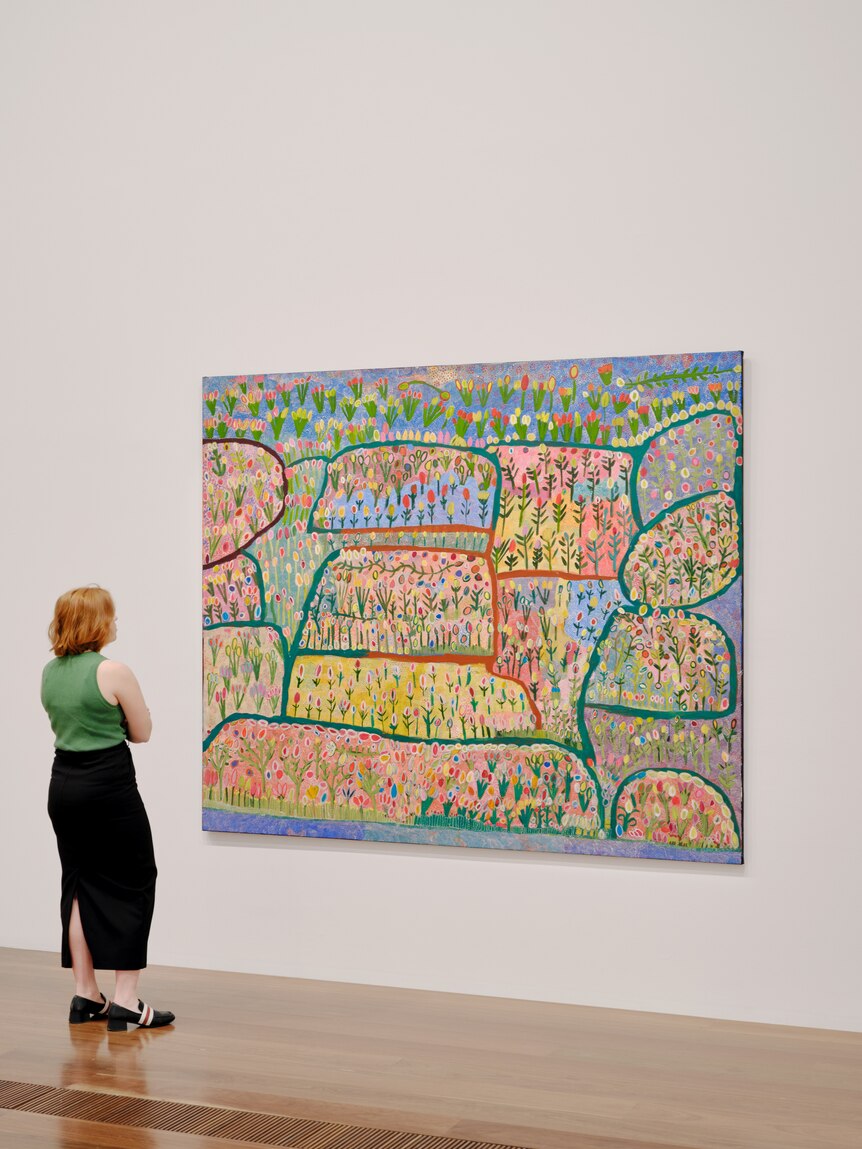 A woman in a green top views a colourful landscape painting by Gwenneth Blitner on display in Wurrdha Marra at NGV Australia.