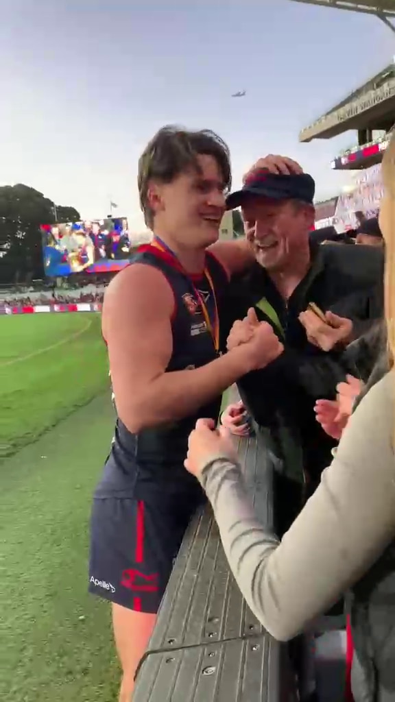 A young man in AFL gear and a man smile and put an arm around each other at the boundary fence of an AFL oval.