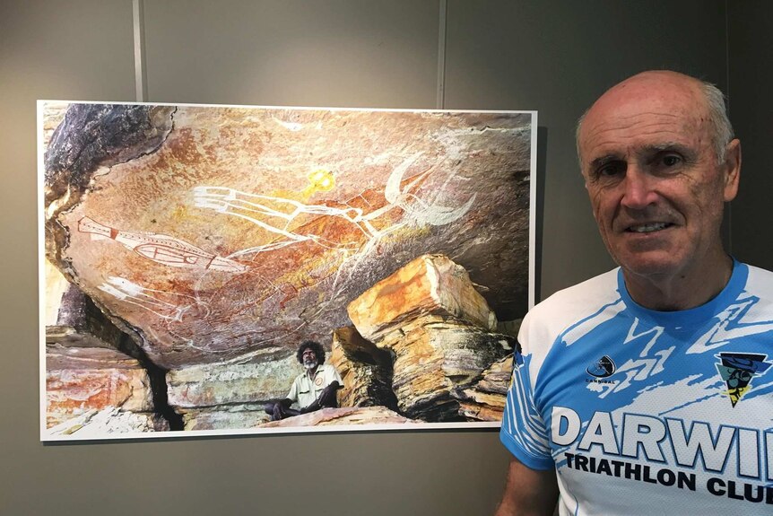David Hancock stands in a triathlon shirt next to a photograph of an Indigenous man sitting under rock art