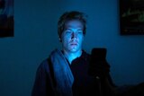 Isaac Freeman sitting in a dark bedroom with the blue light from his phone illuminating his face.
