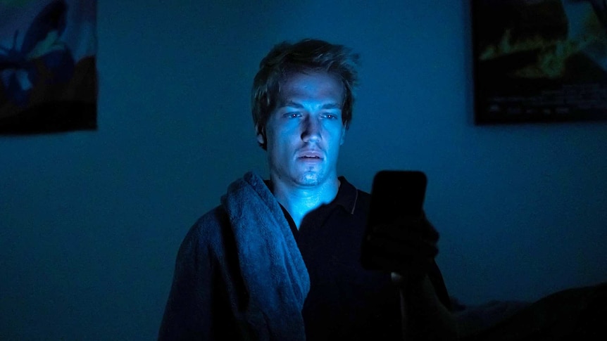 Isaac Freeman sitting in a dark bedroom with the blue light from his phone illuminating his face.