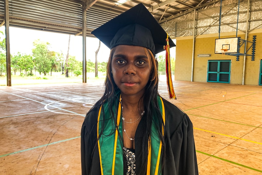 A young indigenous woman stares at the camera, wearing a graduation frock.
