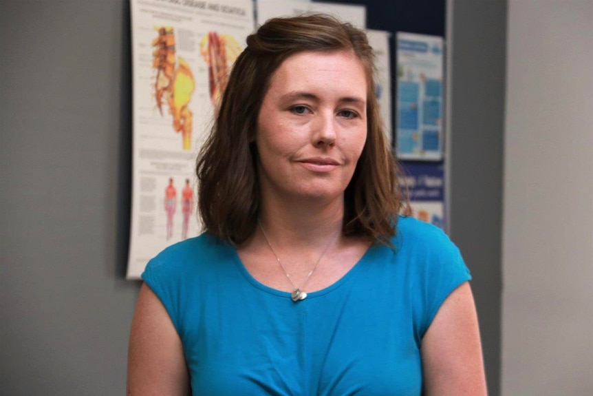 A mid shot of a middle aged woman with brown hair and a blue top standing inside a medical centre posing for a photo.