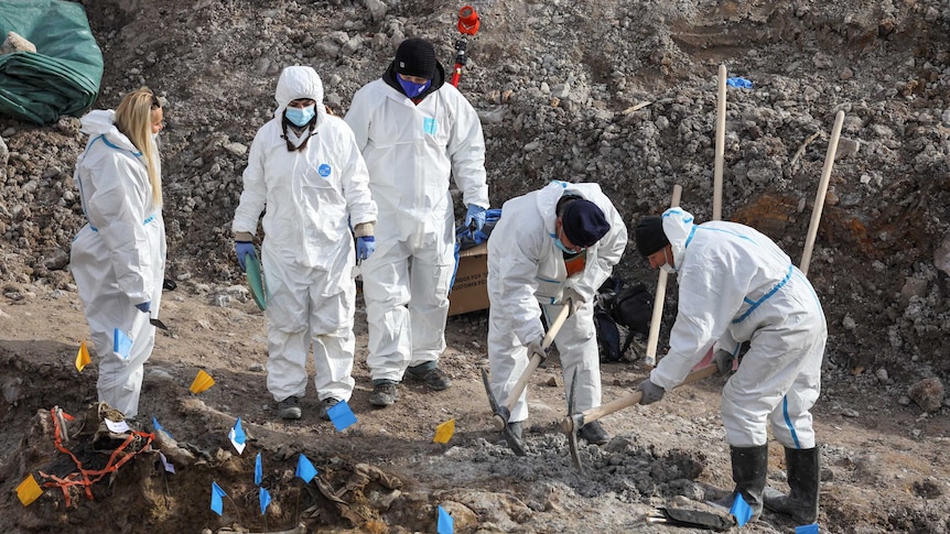 There are five people in white protective hazmat suits looking through dirt, with two on the right using pick-axes.
