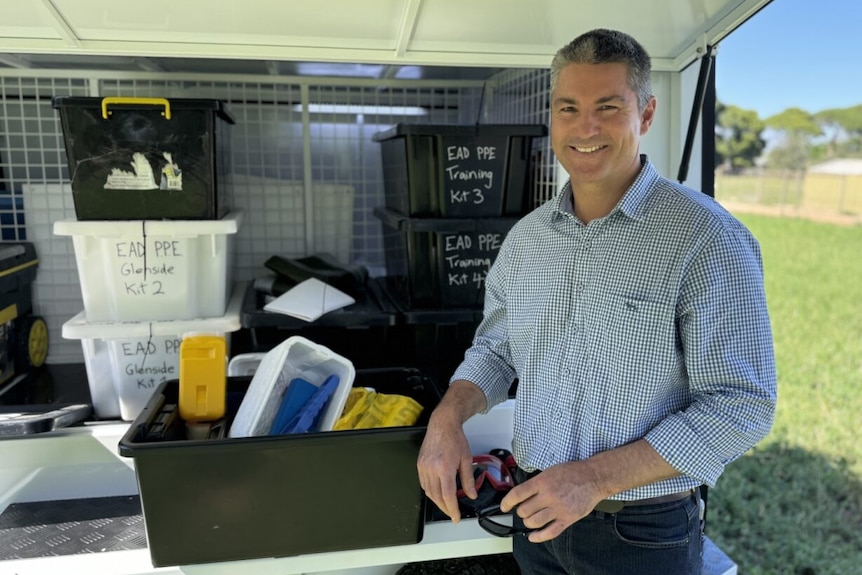 A smiling man with short, grey hair stands in front of a trailer full of veterinary supplies.