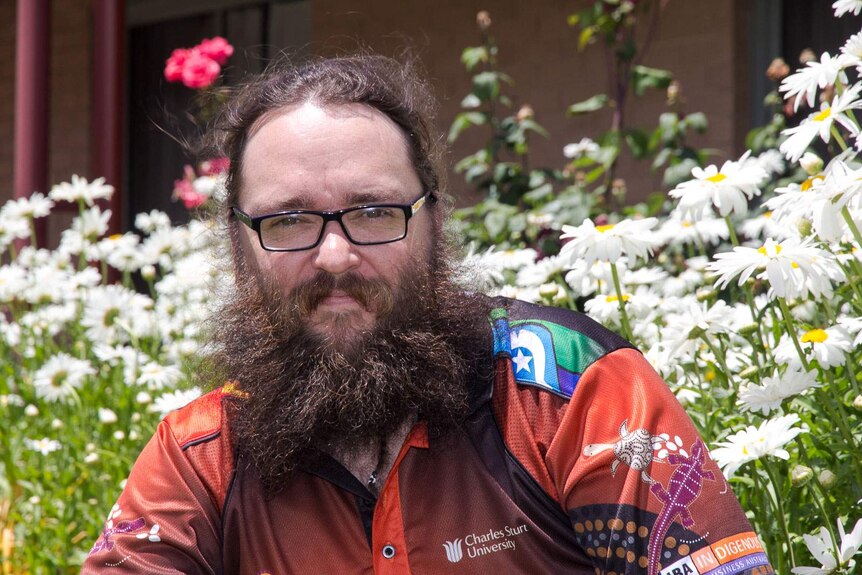 A man with a beard wearing a Charles Sturt University indigenous shirt in front of flowers