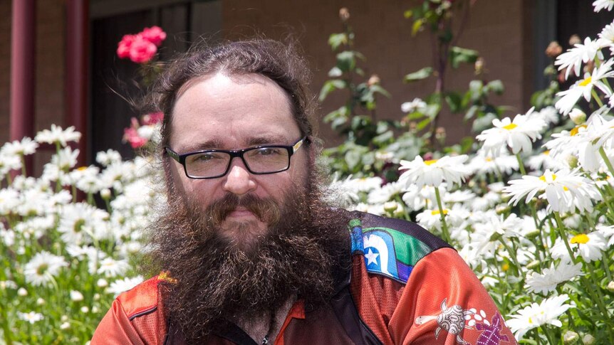 A man with a beard wearing a Charles Sturt University indigenous shirt in front of flowers