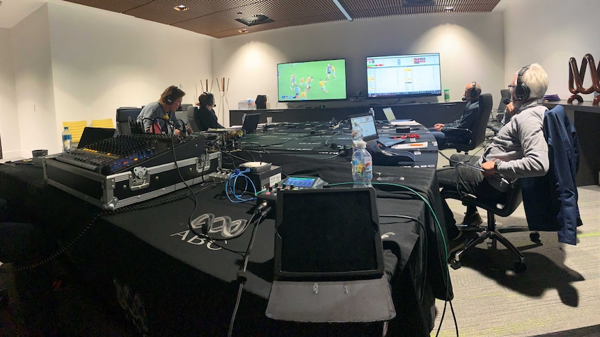 A group of ABC broadcasters sit around a table with headphones on, calling an AFL game off TV.