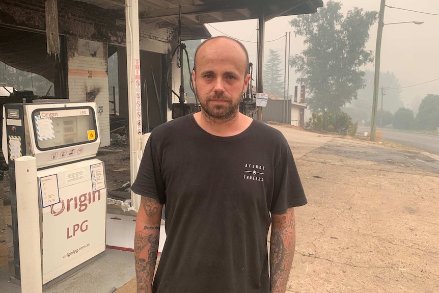 A man stands in front of a burnt out petrol station.