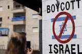A white wall shows a boycott israel stencil with a red mark over the israeli flag