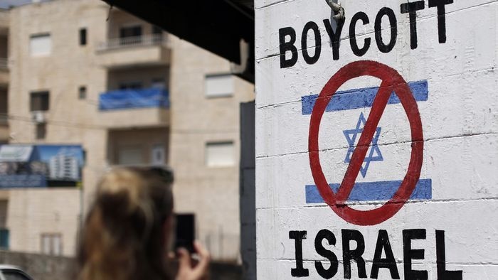 A white wall shows a boycott israel stencil with a red mark over the israeli flag