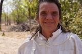 A woman with dark hair and wearing a white beekeeping suit smiles at the camera.