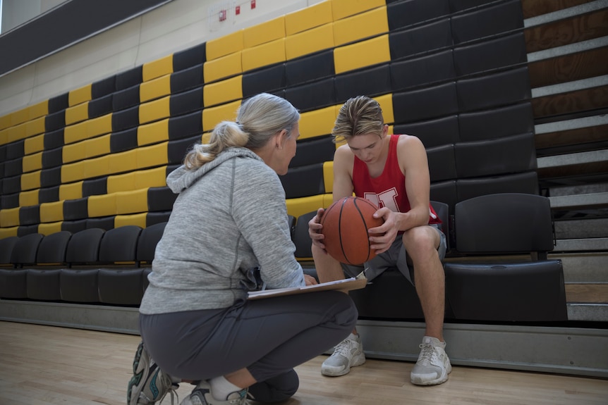 Teenage boy in a red singlet holding a basketball, sitting down and looking sad, being coached by a woman with grey hair.