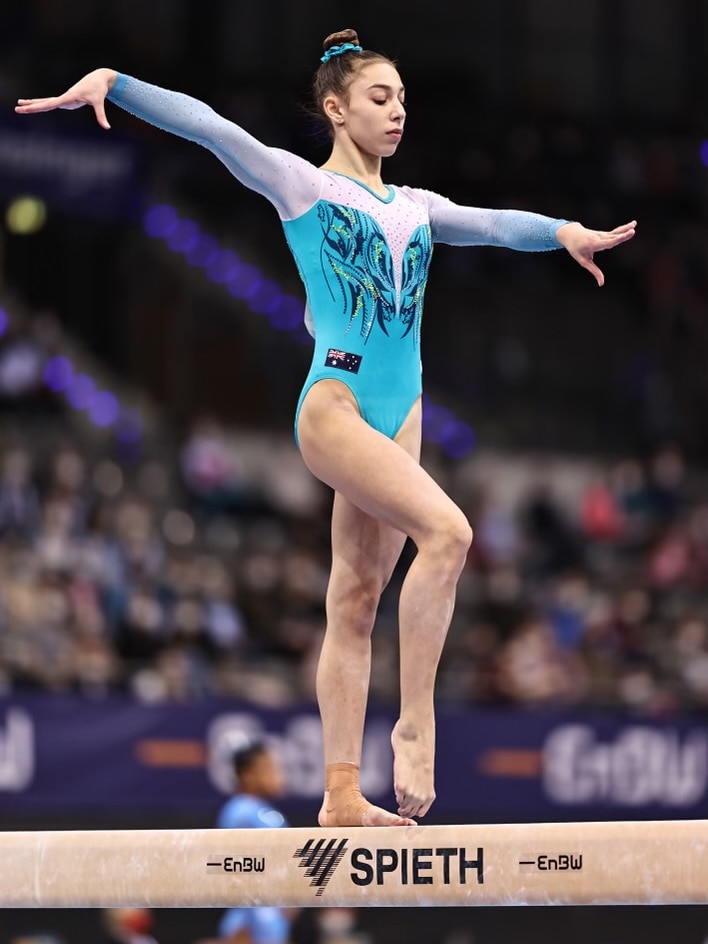 A female gymnast stands on the balance beam with her arms to out and fingers flexed during a routine.