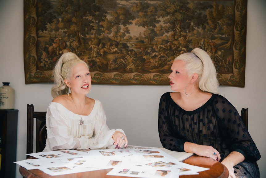 Two platinum blonde women wearing semi-sheer dresses sit at a table discussing designs.
