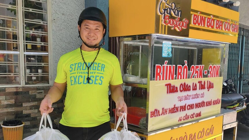A man in yellow t-shirt holding plastic takeaway bags outside a street noodle stall. 
