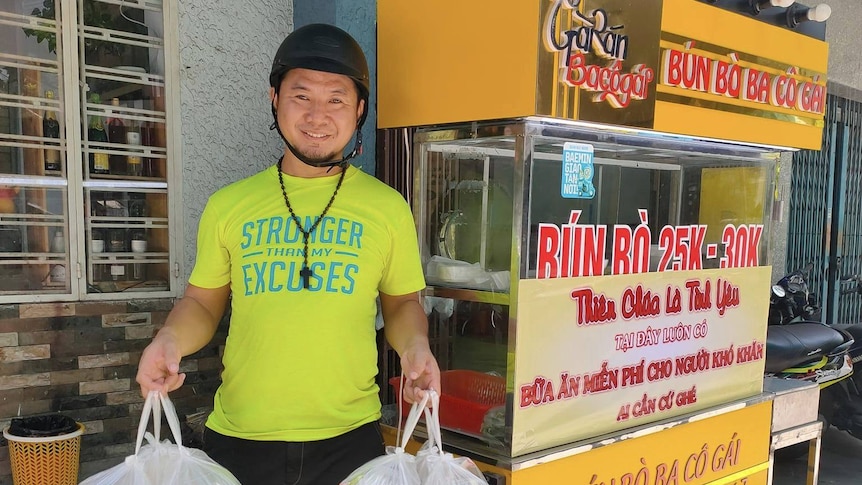 A man in yellow t-shirt holding plastic takeaway bags outside a street noodle stall. 