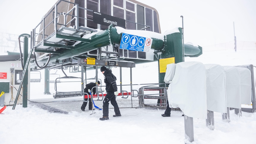 Two people in black work on a chairlift with snow on the ground