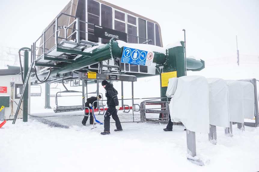 Two people in black work on a chairlift with snow on the ground