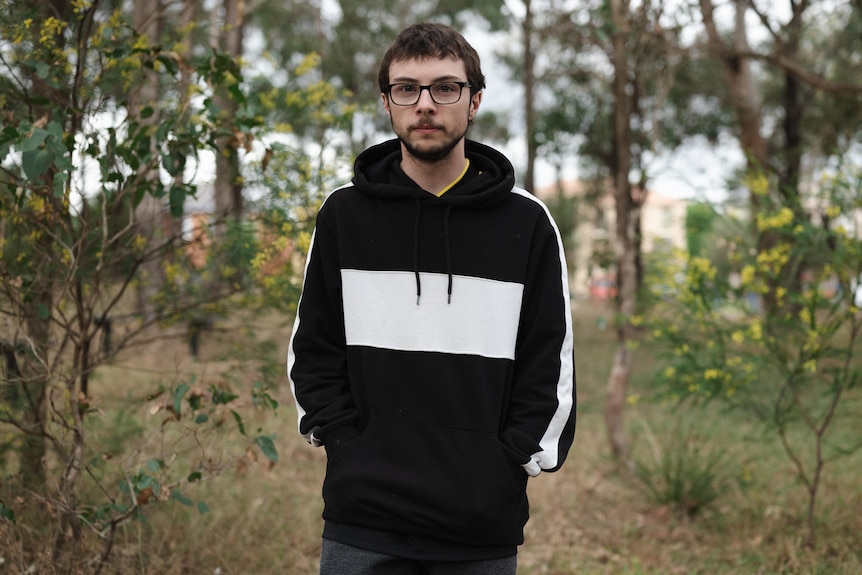 A teenager wearing a hoodie stands in a bushland park his hands in his pockets. He looks at the camera with a neutral expression