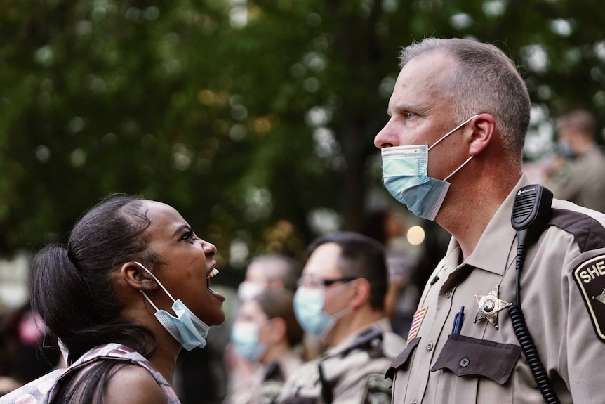 A black woman is seen on the left with a surgical mask pulled down as she yells at a deputy who stares above her.
