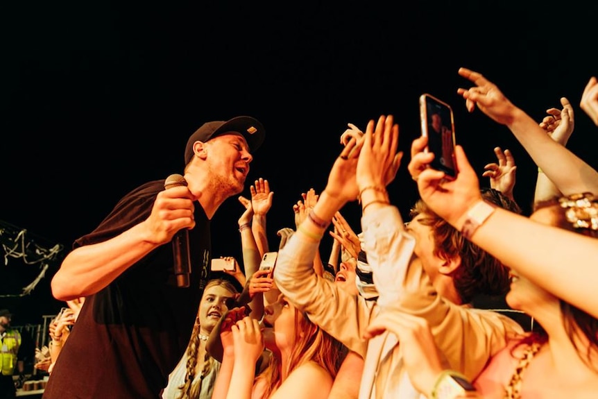 A rapper leans into a cheering crowd, holding a microphone.
