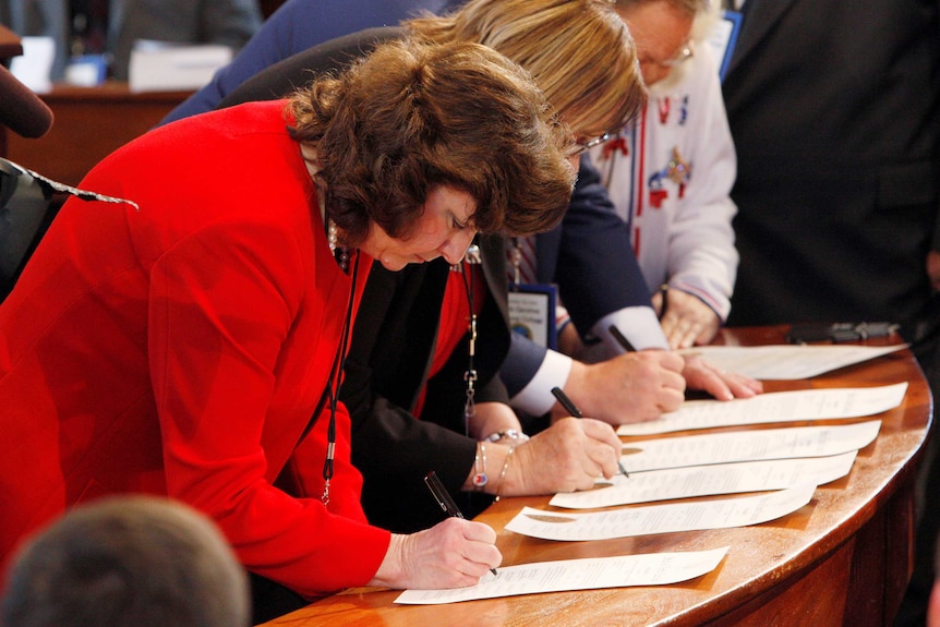 A group of people lean over a wooden desk to sign a certificate