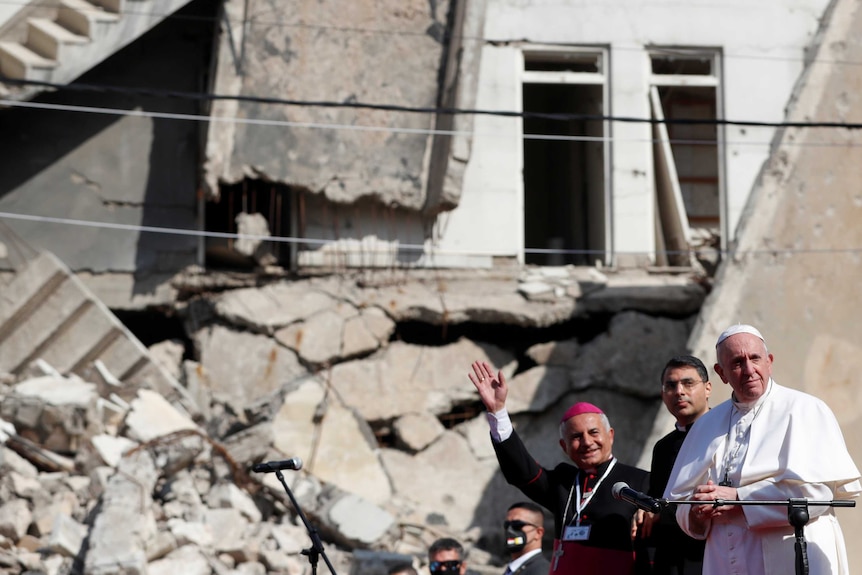 Le pape se tient aux côtés de deux religieux irakiens devant un bâtiment endommagé par la guerre par une journée ensoleillée.