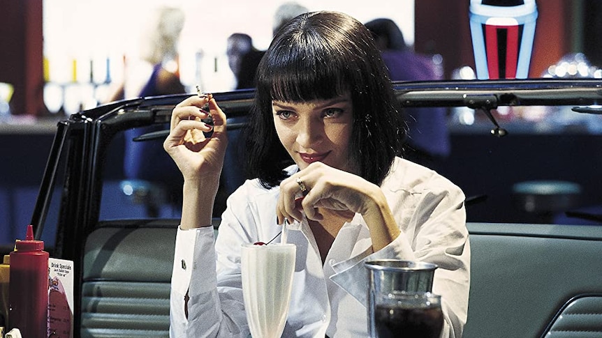 A woman with short black hair and a short fringe sits at a retro-style diner booth drinking a vanilla milkshake 