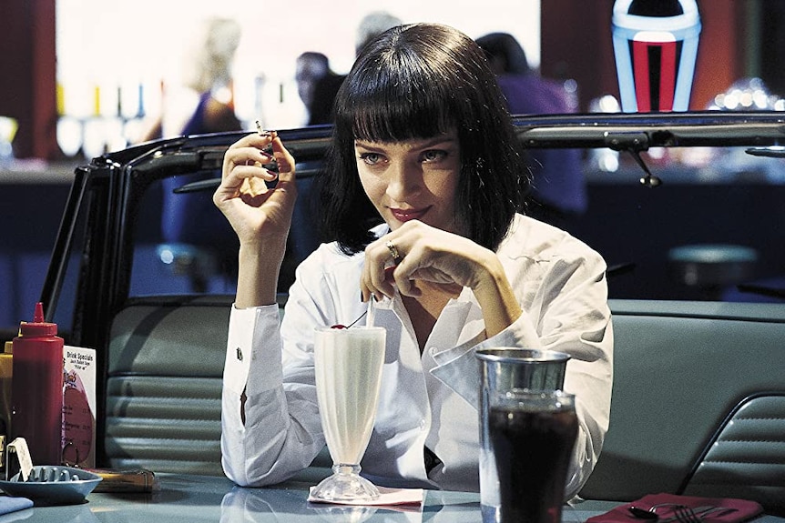 A woman with short black hair and a short fringe sits at a retro-style diner booth drinking a vanilla milkshake 